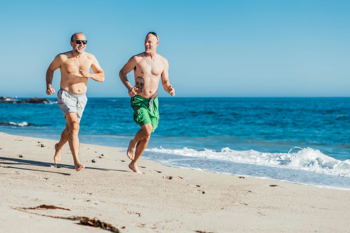 Two men in their 50s running together on an open beach — patients who arrive in their seventies still strong, alert, and engaged with life