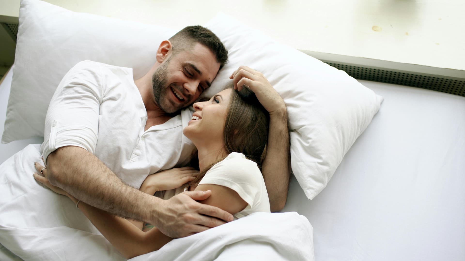Mid-life couple in bed laughing together, both in white tops — restored intimacy when ED's underlying cause is properly diagnosed and treated
